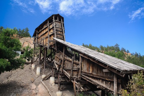 Abandoned wooden structure on a hillside, surrounded by trees and a blue sky.