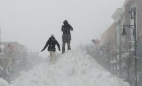Two people stand on a large snow pile in a snowy urban area, with snow falling heavily around them.