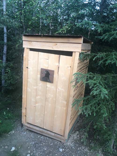 A wooden outhouse with a slanted roof, set among trees in a natural outdoor environment.