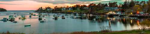 A serene coastal scene at sunset, featuring boats in a calm harbor surrounded by trees and quaint houses.