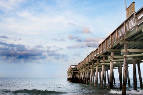 A wooden pier extends over calm waters under a cloudy sky, with light posts lining the walkway.