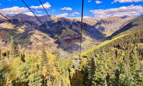 A scenic view from a cable car, overlooking a valley surrounded by mountains and vibrant autumn foliage.