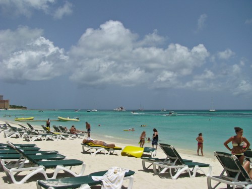 A sunny beach scene with people relaxing on lounge chairs, swimming, and boats in the turquoise water under a cloudy sky.
