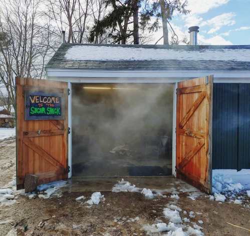 A wooden shed with open doors, emitting steam, surrounded by snow and trees, with a sign reading "Welcome to the Sugar Shack."