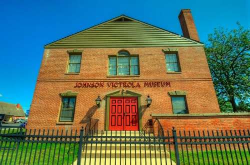Brick building with a sloped roof, red doors, and "Johnson Victrola Museum" sign, surrounded by a black fence.