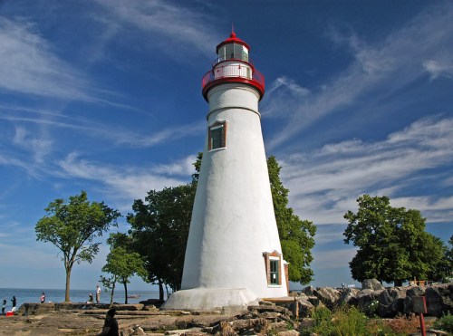 A white lighthouse with a red top stands by the shore, surrounded by trees and a blue sky with wispy clouds.