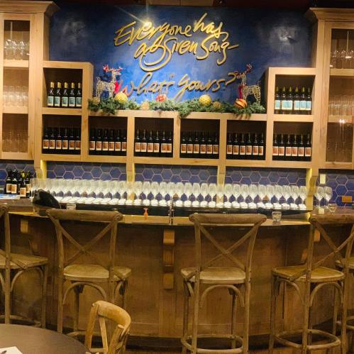 A stylish bar with a blue wall, shelves of wine bottles, and a row of glasses on the counter. Decorated with a quote.