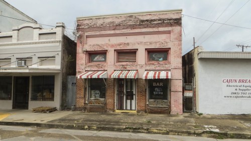 A weathered pink building with a striped awning and "BAR" sign, surrounded by other storefronts on a quiet street.