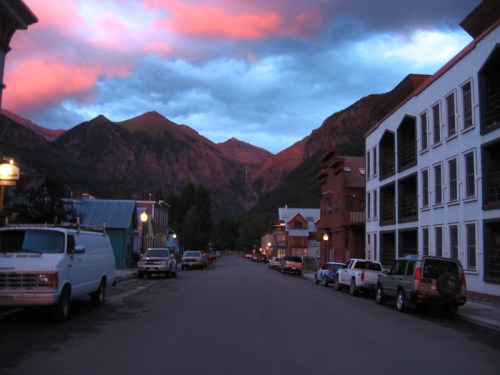 A quiet mountain street at dusk, with colorful clouds and buildings lining the road, surrounded by towering peaks.