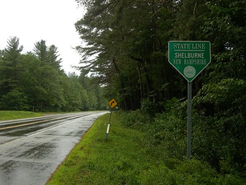 Green state line sign for Shelburne, New Hampshire, beside a wet road lined with trees on a rainy day.