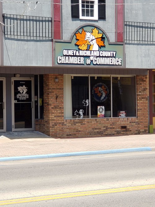 Sign for the Olney & Richland County Chamber of Commerce on a brick building, with a cat logo and storefront windows.