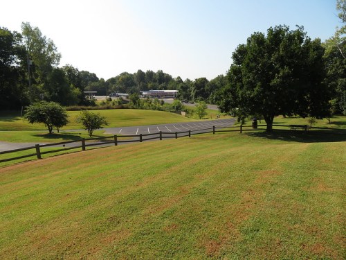 A grassy landscape with trees, a parking lot, and a distant building under a clear blue sky.