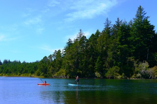 A person paddleboarding and another in a kayak on a calm lake, surrounded by lush green trees and a clear blue sky.
