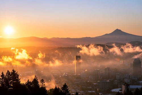 Sunrise over a cityscape with fog, featuring distant mountains and a prominent peak in the background.