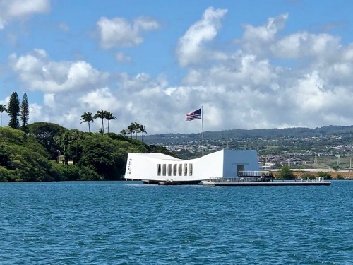 The USS Arizona Memorial, a white structure on water, surrounded by trees and mountains under a blue sky with clouds.