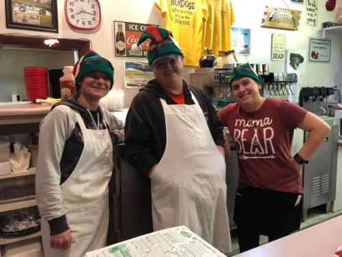 Three smiling employees wearing green elf hats and aprons stand behind a counter in a cozy diner.