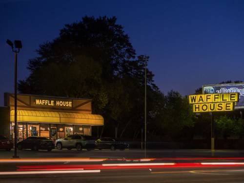 A Waffle House restaurant illuminated at night, with a glowing sign and light trails from passing cars.