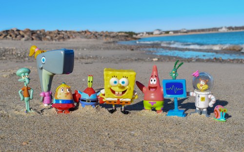 A group of colorful SpongeBob SquarePants toys stands on a sandy beach, with the ocean and rocks in the background.