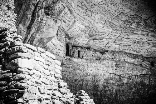 Black and white image of ancient stone ruins beneath a rocky overhang, showcasing weathered textures and structures.