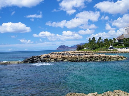 A rocky shoreline extends into clear blue water, with palm trees and mountains in the background under a partly cloudy sky.