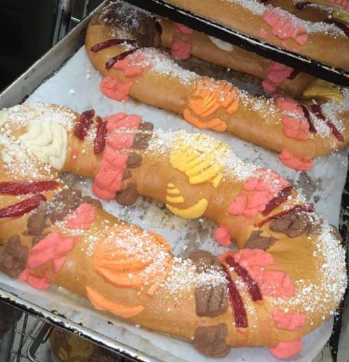 A close-up of colorful sweet bread, decorated with icing and powdered sugar, arranged on a baking tray.
