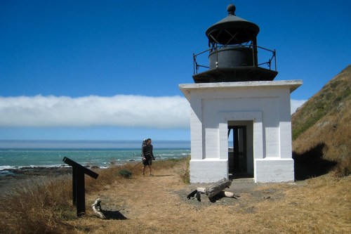 A lighthouse stands by the coast, with a person walking nearby against a backdrop of blue sky and ocean.