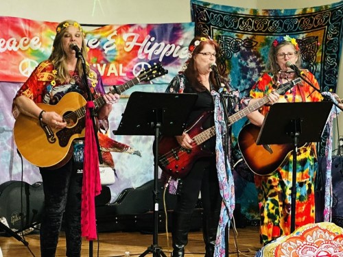 Three women in colorful outfits perform with guitars on stage, surrounded by vibrant, tie-dye backdrops.