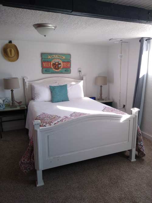 Cozy bedroom with a white bed, colorful bedding, lamps, and vintage milk sign on the wall. Natural light from a window.