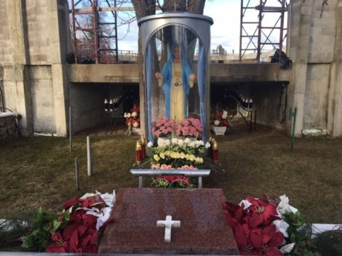 A statue of a saint enclosed in glass, surrounded by flowers and candles at a memorial site.