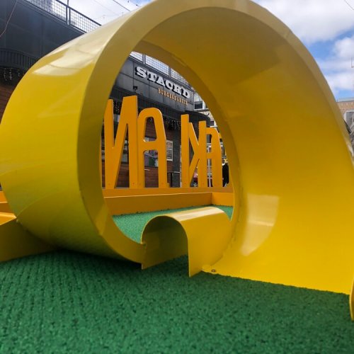 A bright yellow sculpture with the word "KINA" partially visible, set against a green surface and blue sky.