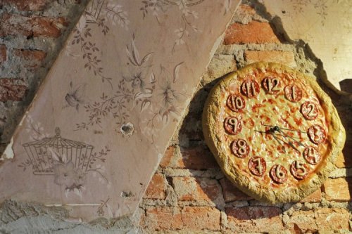 A unique wall clock shaped like a pizza, mounted on a textured brick wall with floral wallpaper.