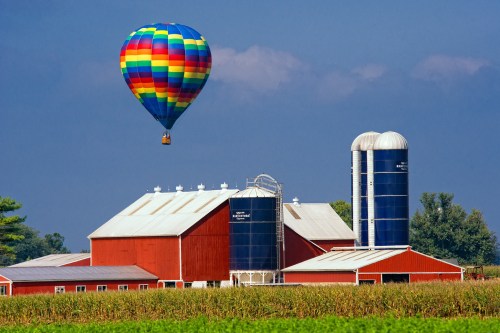 A colorful hot air balloon floats above a red barn and silos in a rural landscape with cornfields.