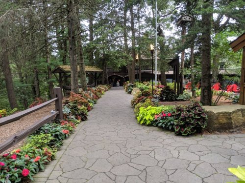 A stone pathway lined with colorful flowers leads through a wooded area to a rustic building in the background.