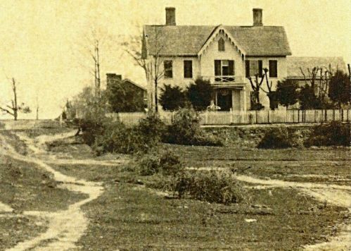 A vintage photograph of a large house surrounded by trees and a dirt path leading up to it.