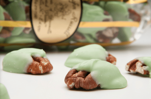 A close-up of chocolate-covered pecans with a green coating, scattered near a decorative jar.