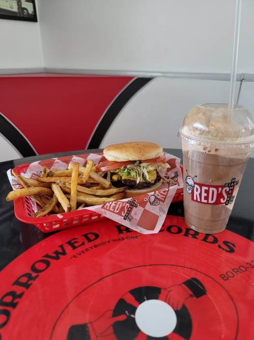 A burger with lettuce and fries in a red basket, alongside a chocolate milkshake on a table with a red design.