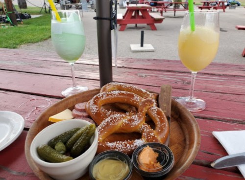 A wooden platter with a large pretzel, pickles, and dipping sauces, alongside two colorful drinks on a picnic table.