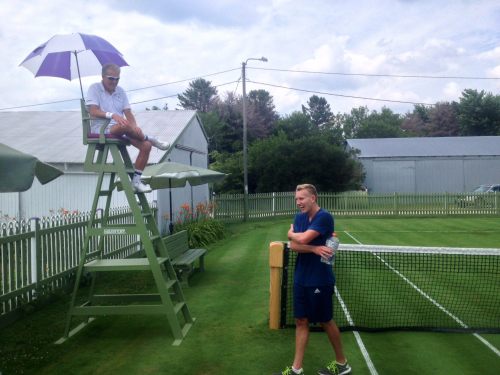 A tennis match in progress with a player sitting on a high chair under an umbrella, while another player stands nearby.