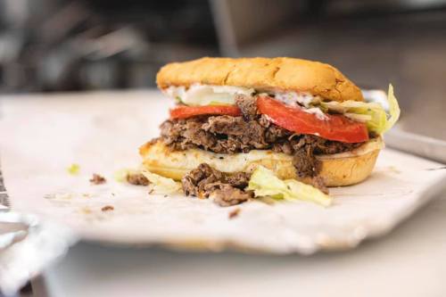 A close-up of a sandwich with sliced beef, lettuce, and tomato on a toasted bun, resting on parchment paper.