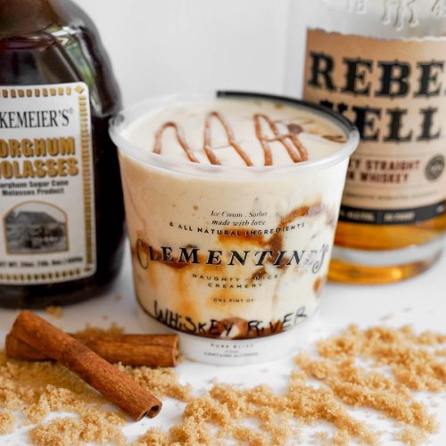 A container of Clementine's whiskey river ice cream, surrounded by whiskey, molasses, cinnamon sticks, and brown sugar.
