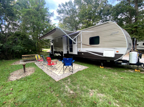 A parked camper trailer with outdoor seating and a rug, surrounded by trees in a grassy area.