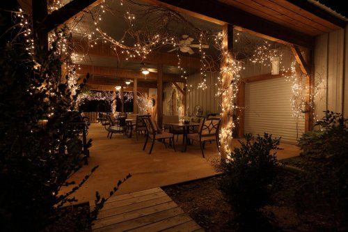 A cozy outdoor space with string lights, tables, and chairs, surrounded by greenery in a warm, inviting atmosphere.
