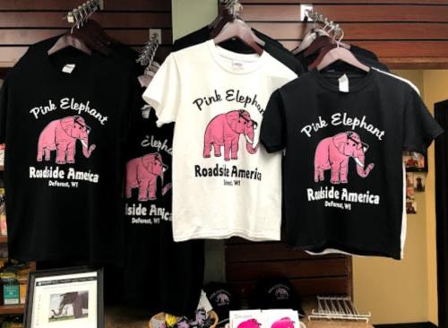 A display of t-shirts featuring a pink elephant design, labeled "Pink Elephant Roadside America," in black and white.
