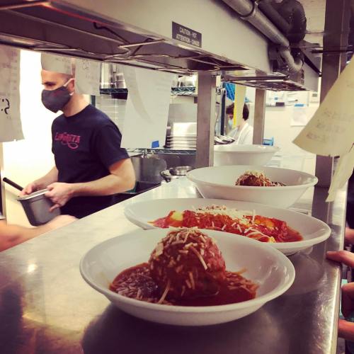 A kitchen scene with bowls of spaghetti and meatballs on a counter, while a masked chef prepares food in the background.