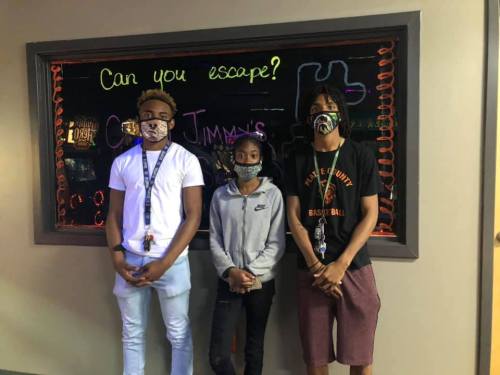Three young people wearing masks stand in front of a colorful "Can you escape?" sign at an escape room venue.