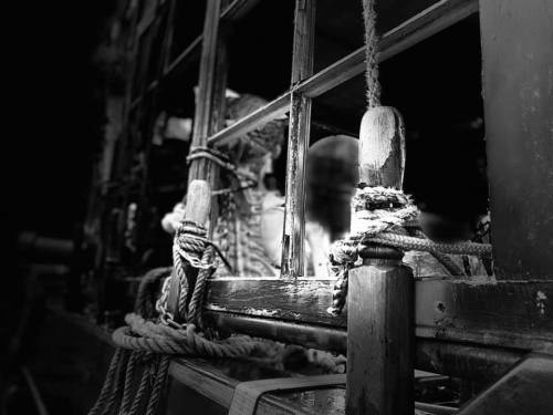 Close-up of a wooden ship's railing with ropes, captured in black and white, highlighting textures and shadows.