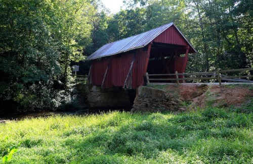 A red covered bridge stands over a stream, surrounded by lush greenery and trees under a clear blue sky.