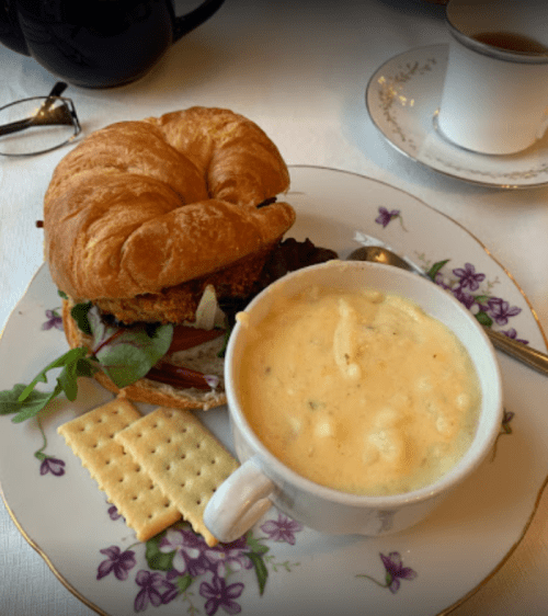 A croissant sandwich with greens and a cup of creamy soup, served with crackers on a floral plate.