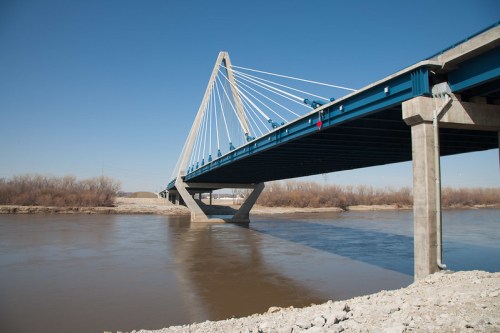 A modern cable-stayed bridge spans a river under a clear blue sky, with rocky banks visible on either side.