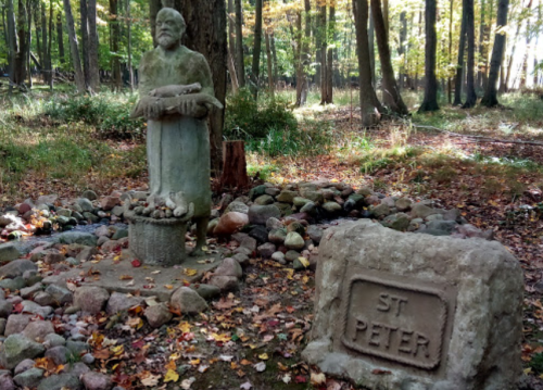 A stone statue of St. Peter stands in a forest clearing, surrounded by rocks and autumn leaves.